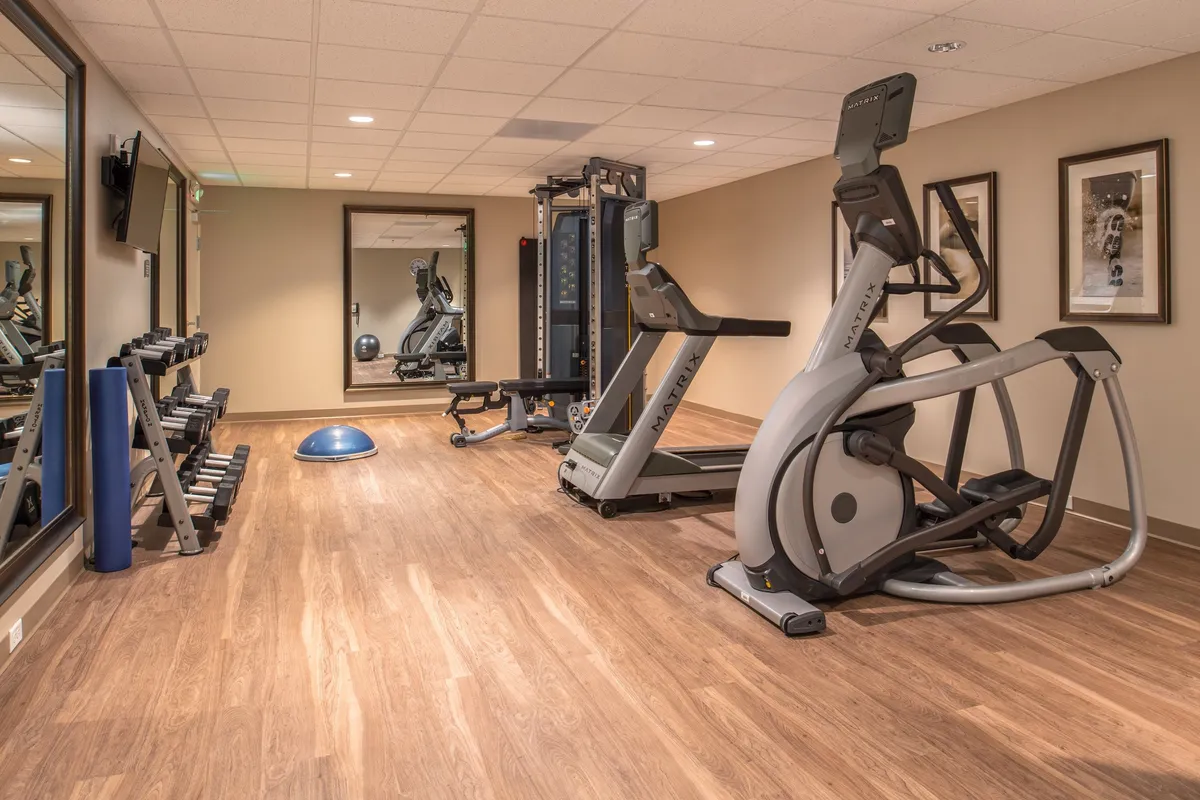 This dynamic fitness center image includes an elliptical machine, weight rack, exercise mat, and expansive fitness mirrors. The clean, well-lit space conveys a motivating and active vibe, highlighting a range of workout opportunities.