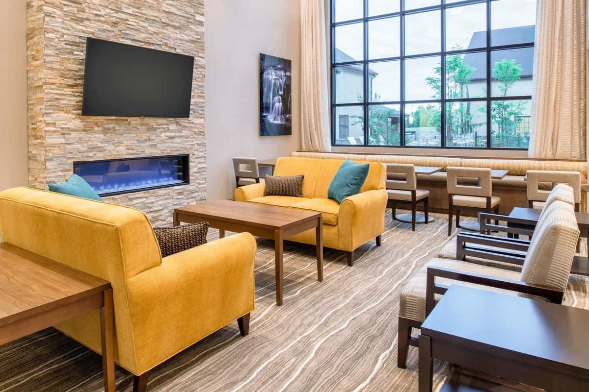 A warmly lit living area featuring plush yellow sofas arranged around a coffee table, with a modern stone feature wall and large windows that let in natural light. Modern chairs add to the room's versatility for social gatherings or quiet relaxation. The space is designed for comfort and offers an inviting ambiance for guests to unwind, watch TV, or enjoy a conversation.