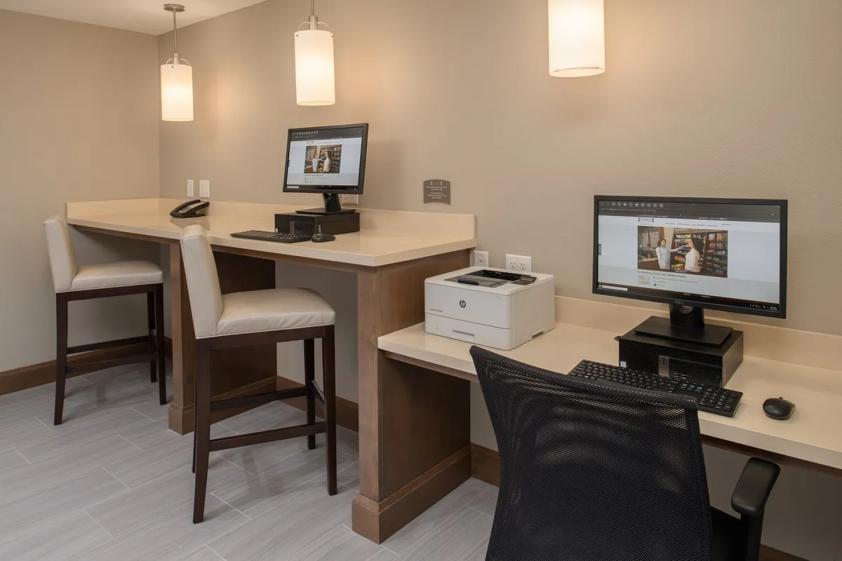 A modern, inviting workspace area featuring computers, bar-style seating, and a printer. The lighting is bright and conducive to productivity, offering guests a place to work, study, or connect online. The atmosphere balances professionalism and comfort, providing a seamless blend of home and hotel perks.