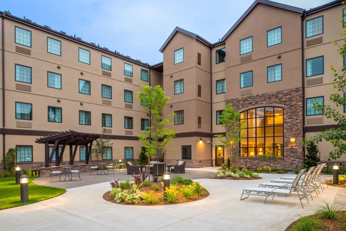 This exterior scene displays a lushly landscaped patio area with a pergola, comfortable seating, and large windows that glow with warm indoor light. The atmosphere is sheltered yet open, hinting at both outdoor relaxation and seamless transition between inside and out.
