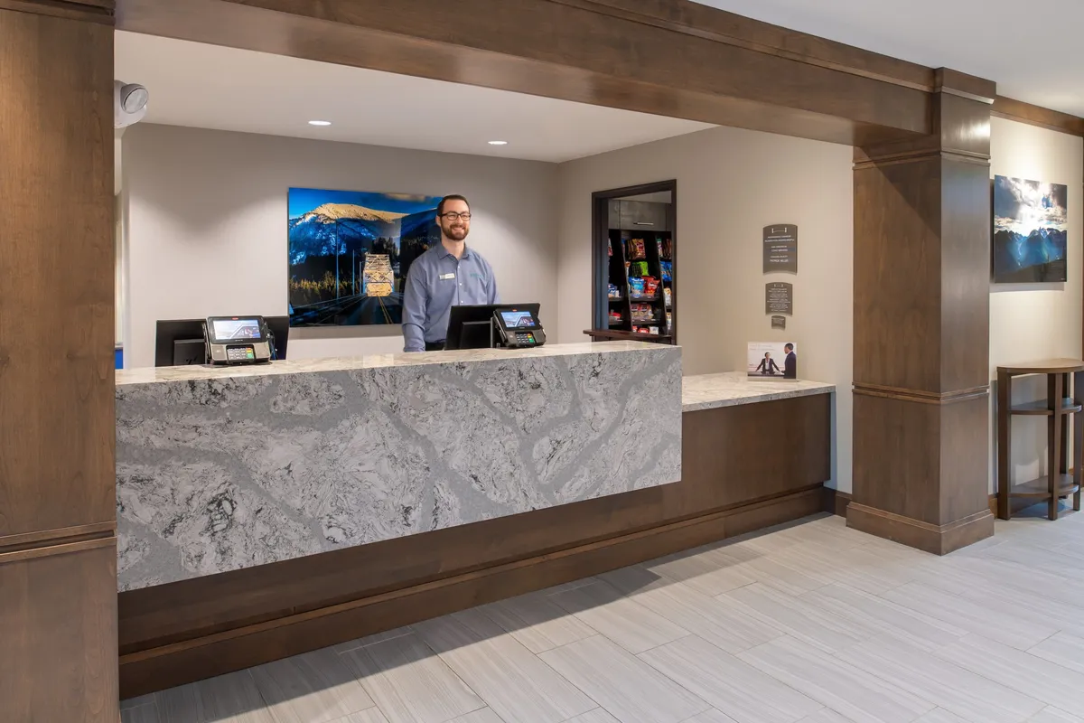 The image captures the reception desk area, where a staff member is assisting guests. Local landscape-inspired artwork decorates the wall, and the atmosphere is warm and welcoming. The setting alludes to the start of a relaxing, well-supported getaway experience.
