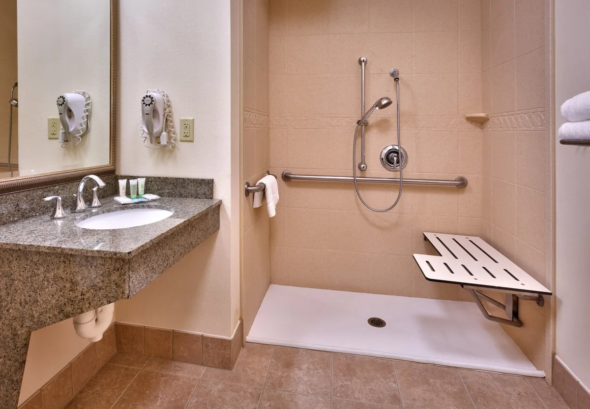 A contemporary bathroom scene with a granite countertop sink, a spacious shower featuring a handheld showerhead, support bars, and a shower bench. Fresh towels are visible, contributing to a sense of cleanliness and luxury, while the accessibility features add a modern, inclusive touch.