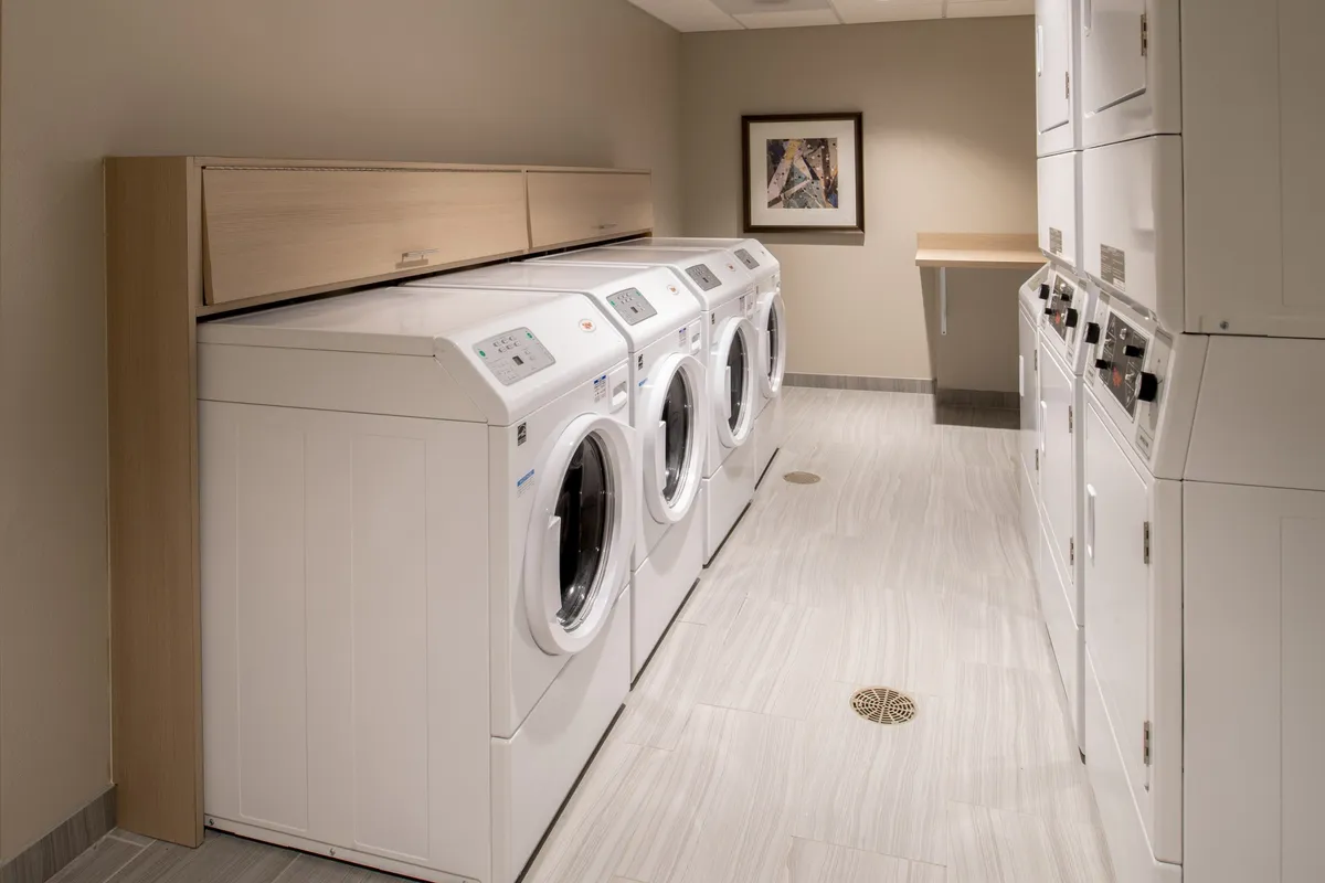 A clean, well-lit laundry area with multiple washing machines, a dryer, and a counter for folding clothes. Framed artwork adds a touch of warmth to the practical space, and the organized layout emphasizes convenience and community use.