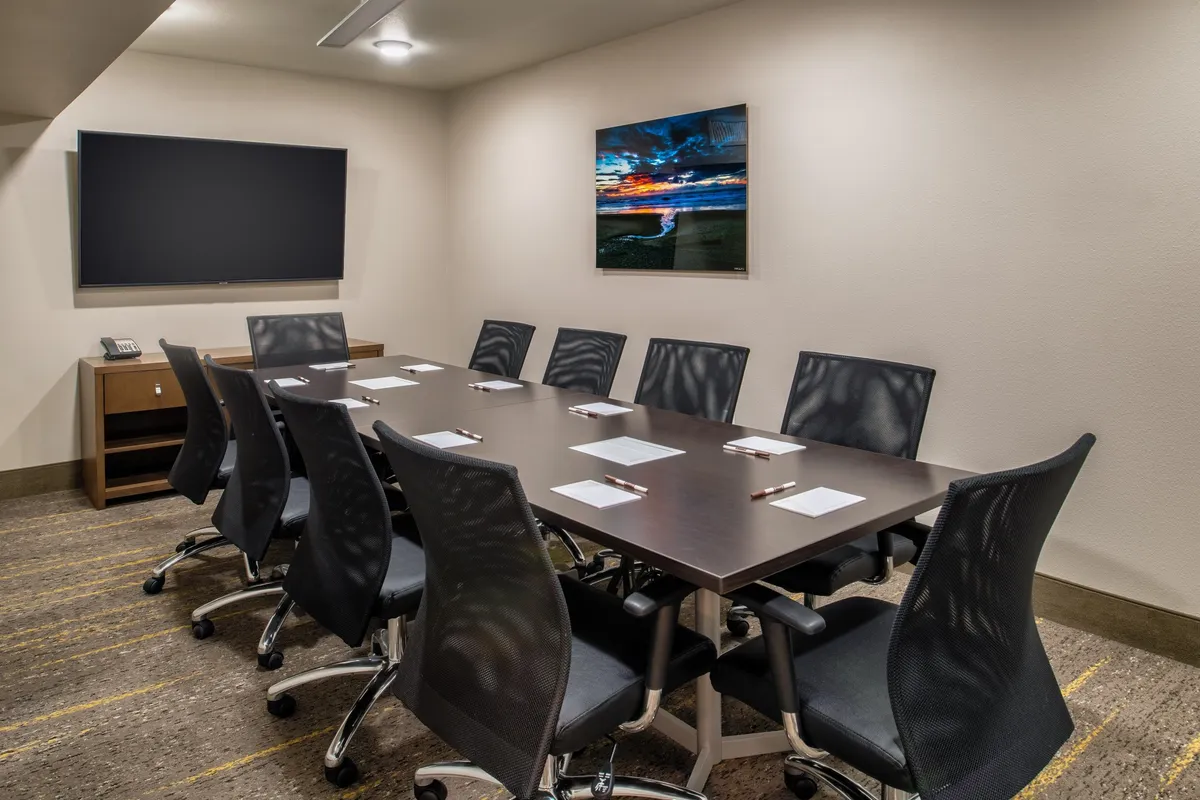 A professional business center with a large meeting table, ergonomic chairs, a television screen for presentations, and tasteful wall art. The setting supports focused work, collaboration, and productivity.