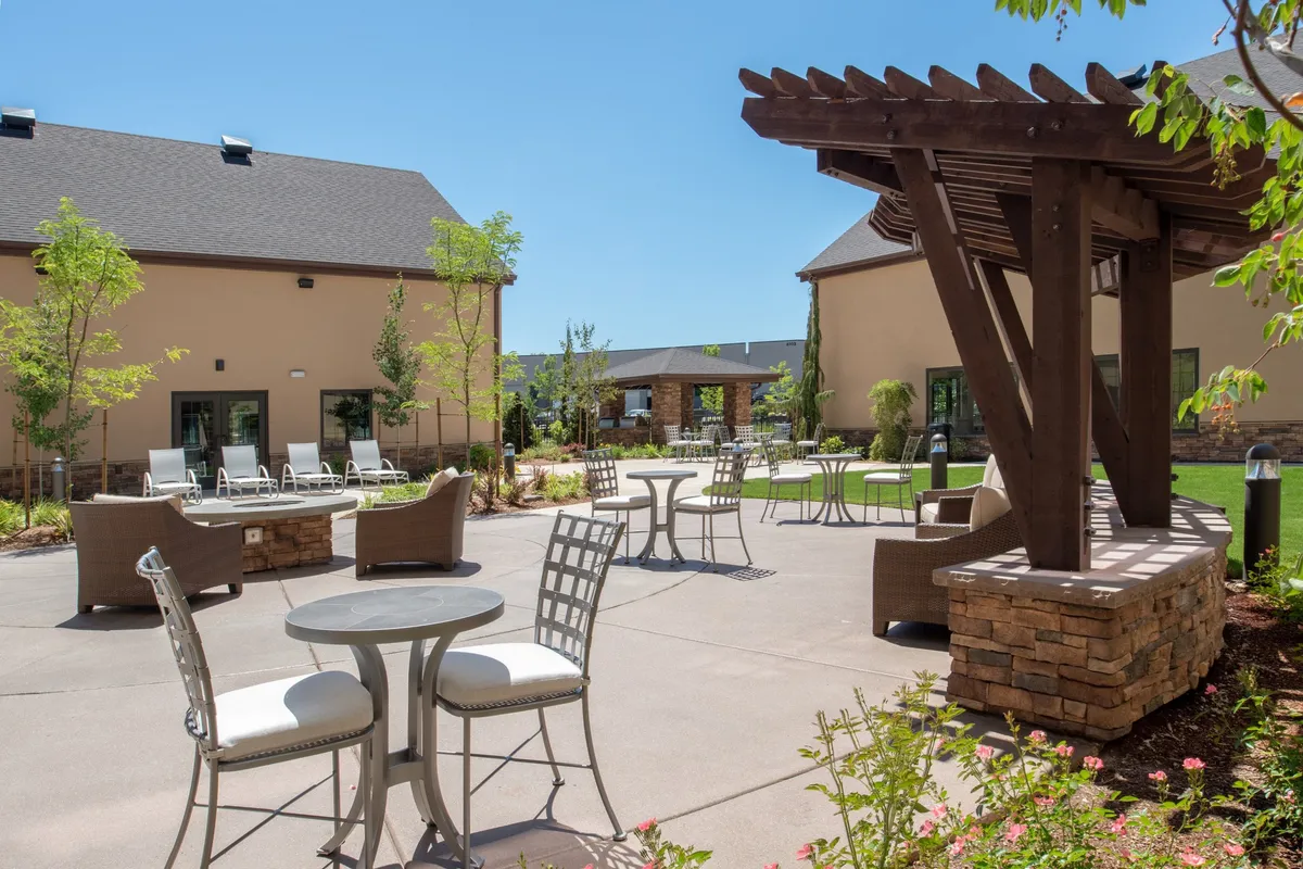 An inviting outdoor seating area under a pergola, surrounded by lush landscaping and stone-accented furniture. The space is arranged for social gatherings, outdoor dining, or simply relaxing with fellow guests. The sunny setting is perfect for barbecues or evening receptions.
